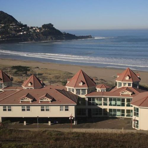 A coastal building complex with red roofs by a sandy beach and calm ocean waves under a clear sky.