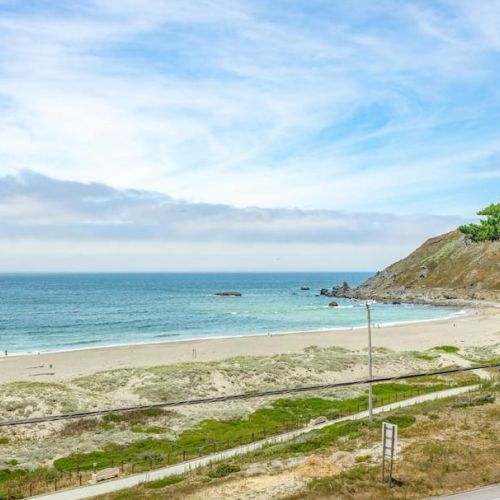 A scenic beach with clear blue water, sandy shoreline, grassy dunes, and a distant rocky hill under a partly cloudy sky.