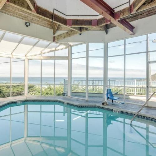 An indoor pool with large windows provides an ocean view, surrounded by a wooden beam ceiling and a single chair on the deck.