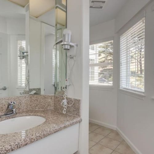 The image depicts a bathroom with a granite countertop, sink, hairdryer, and toiletries, adjacent to a bright window with blinds.