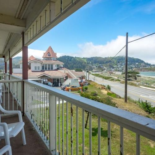 A balcony with chairs overlooks a coastal road, beach, and hills under a blue sky with scattered clouds.