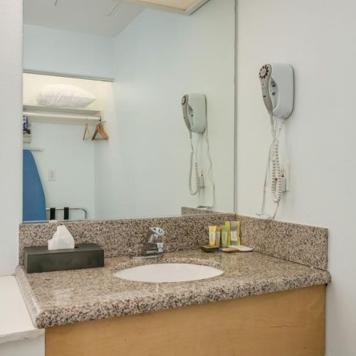 A bathroom countertop with a sink, mirror, hairdryer, toiletries, and a tissue box. There is an ironing board and hangers in the background.