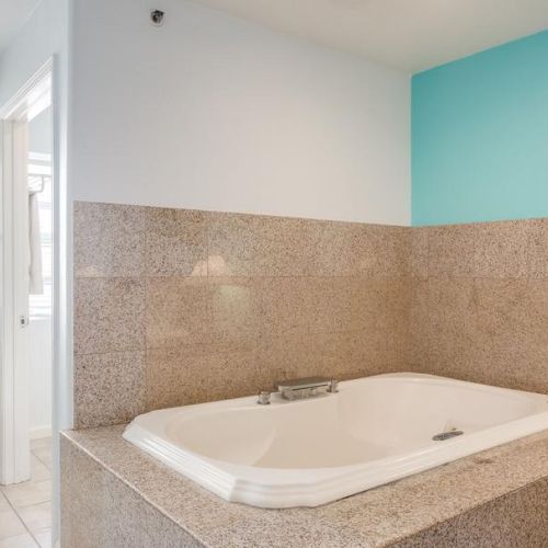 A bathroom with a large, cream-colored bathtub on a tiled platform, set against light blue and white walls with a bright window nearby.