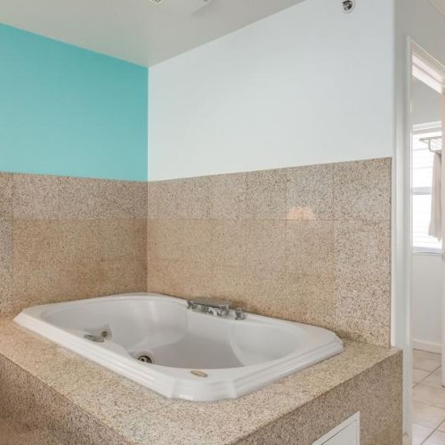 A bathroom features a large, built-in jacuzzi tub with tiled sides, a turquoise and white wall, a wooden nightstand, and natural light from a window.