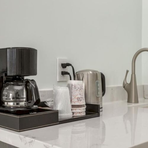 A countertop with a coffee maker, electric kettle, cups, lids, and a faucet on a marble surface.