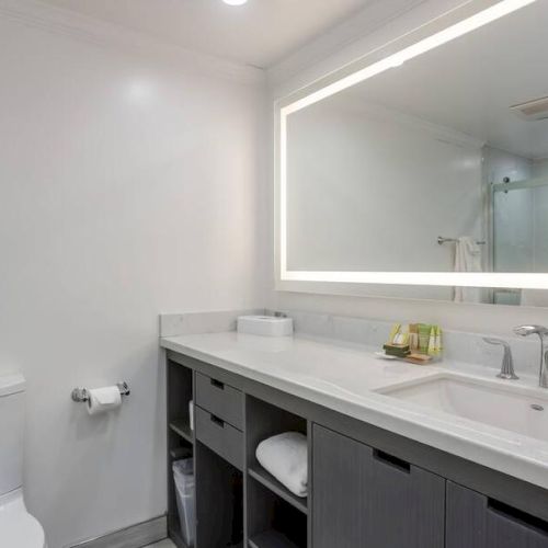 A modern bathroom with a large backlit mirror, sink, and toiletries on the counter, featuring a toilet and a glass shower enclosure.