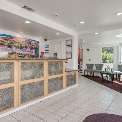 The image shows a brightly lit hotel lobby with a reception desk, chairs in a waiting area, and a colorful painting on the wall.