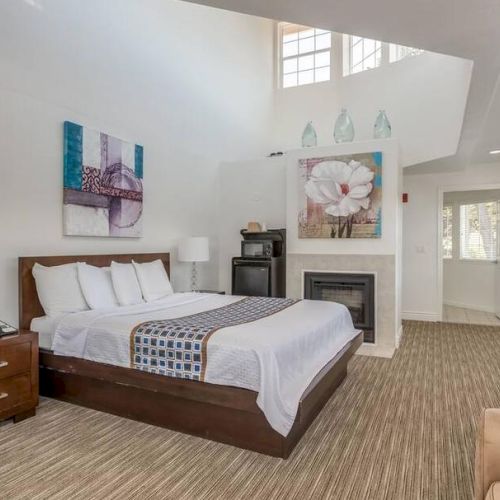 A modern bedroom with a bed, artworks on walls, a fireplace, TV, and seating area. Neutral tones and natural light fill the space.