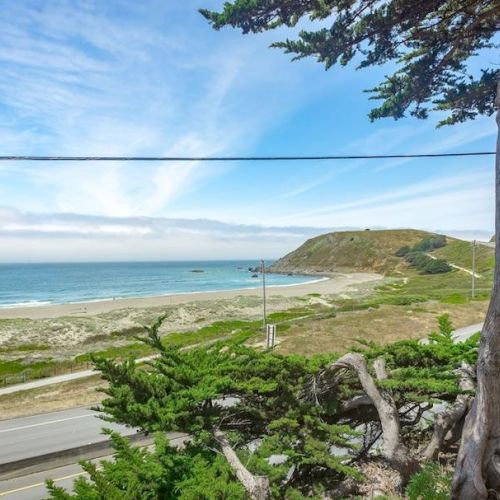 A scenic coastal view with a sandy beach, ocean, hills, and a road lined with trees under a blue sky.