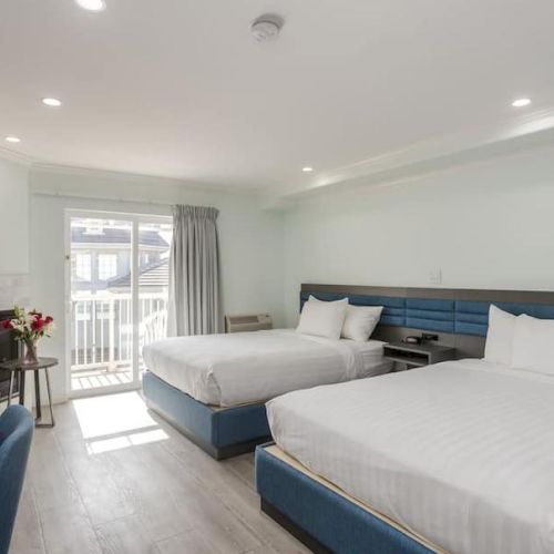 A hotel room with two beds, a desk, and a balcony with a curtain. White walls and bedding, blue headboards. Natural light from windows.