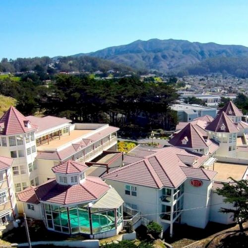 The image shows a complex of large buildings with pointed roofs surrounded by greenery, with mountains visible in the background.