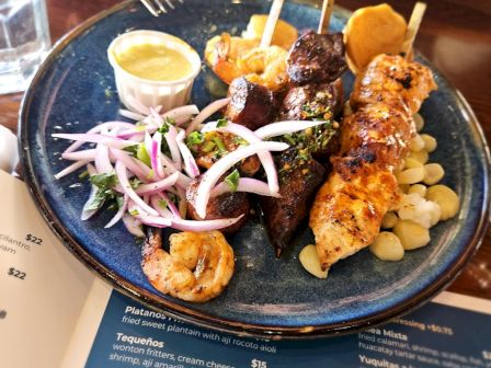 The image shows a plate of skewers with chicken and beef, garnished with onions and sauce, accompanied by shrimp and corn.