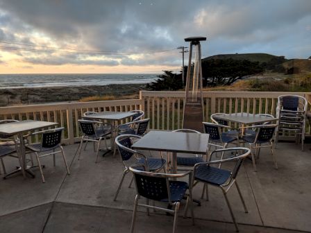 Outdoor patio with metal tables and chairs, overlooking the ocean at sunset. There's a portable heater on the deck.