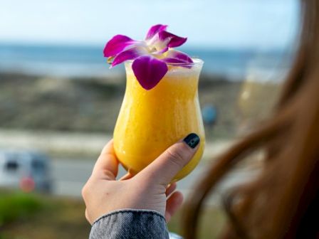 A person holds a yellow drink with a purple flower garnish by the beach, enjoying the ocean view in the background.