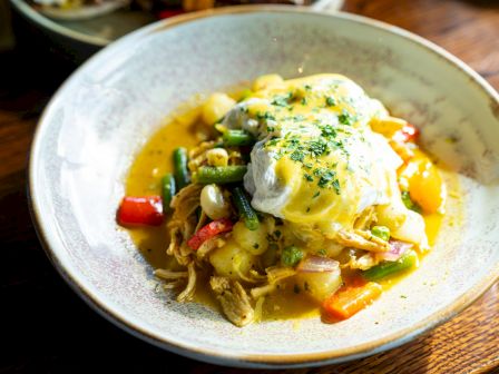 A dish featuring poached eggs on shredded meat with mixed vegetables in a sauce, garnished with herbs, on a ceramic plate.