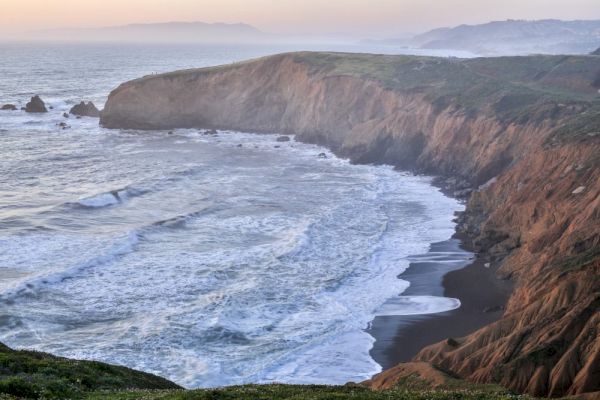 A scenic coastal landscape with cliffs, waves crashing on a sandy beach, and distant hills under a calm sky, creating a serene view.