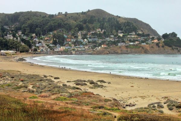 A coastal scene with a sandy beach, ocean waves, and a hillside with houses and trees. Overcast sky adds to the serene atmosphere.