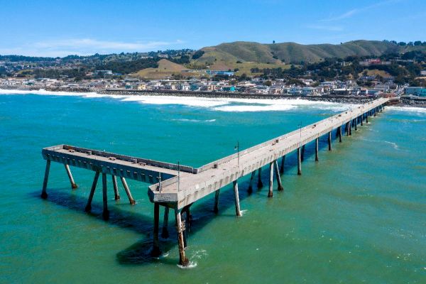 A long pier extends over turquoise water towards a coastal town with hills in the background, under a clear blue sky.