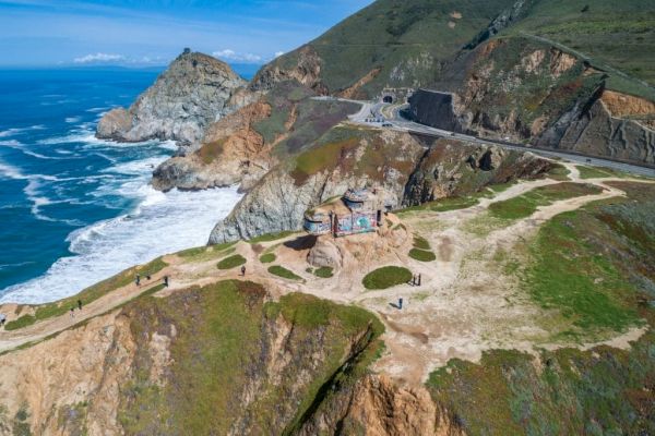 A coastal view shows a scenic overlook above cliffs and ocean waves, with a road leading through the rugged landscape under a blue sky.