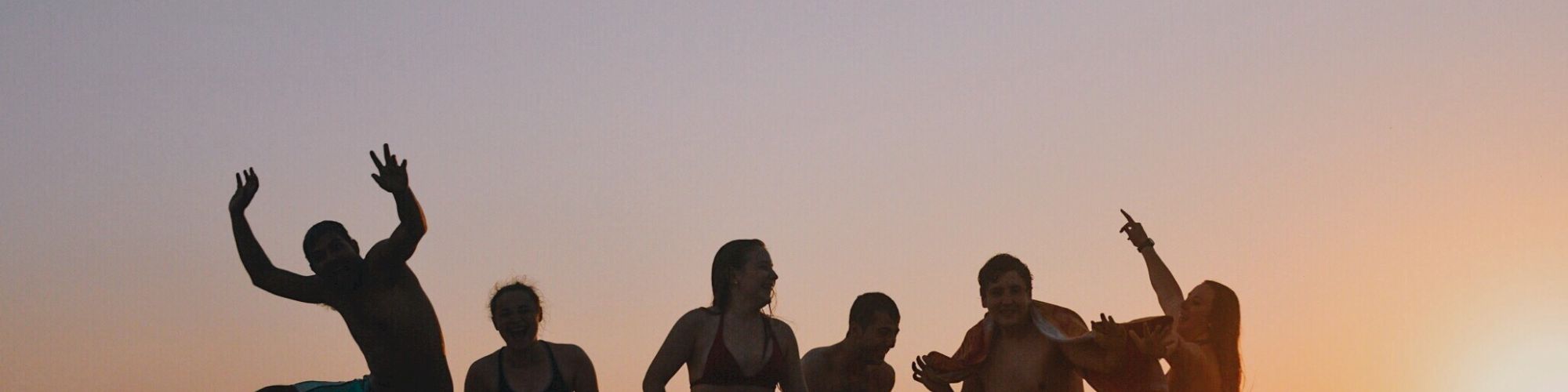 A group of people enjoying sunset on a beach, silhouetted against the colorful sky, with one person playfully jumping.