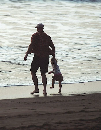 A person and a child are walking along a beach, near the shoreline, with gentle waves in the background.