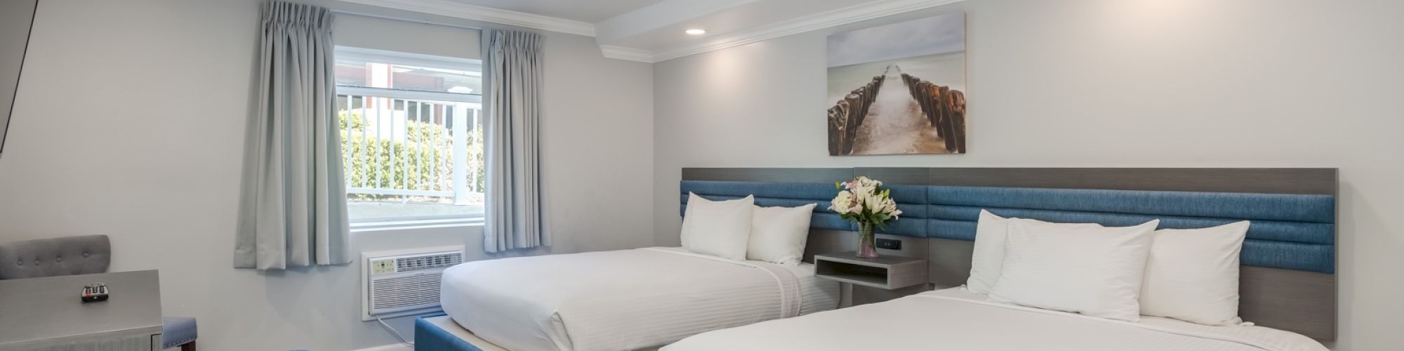 The image shows a modern hotel room with two beds, armchair, desk, and window, decorated in a blue and white color scheme.